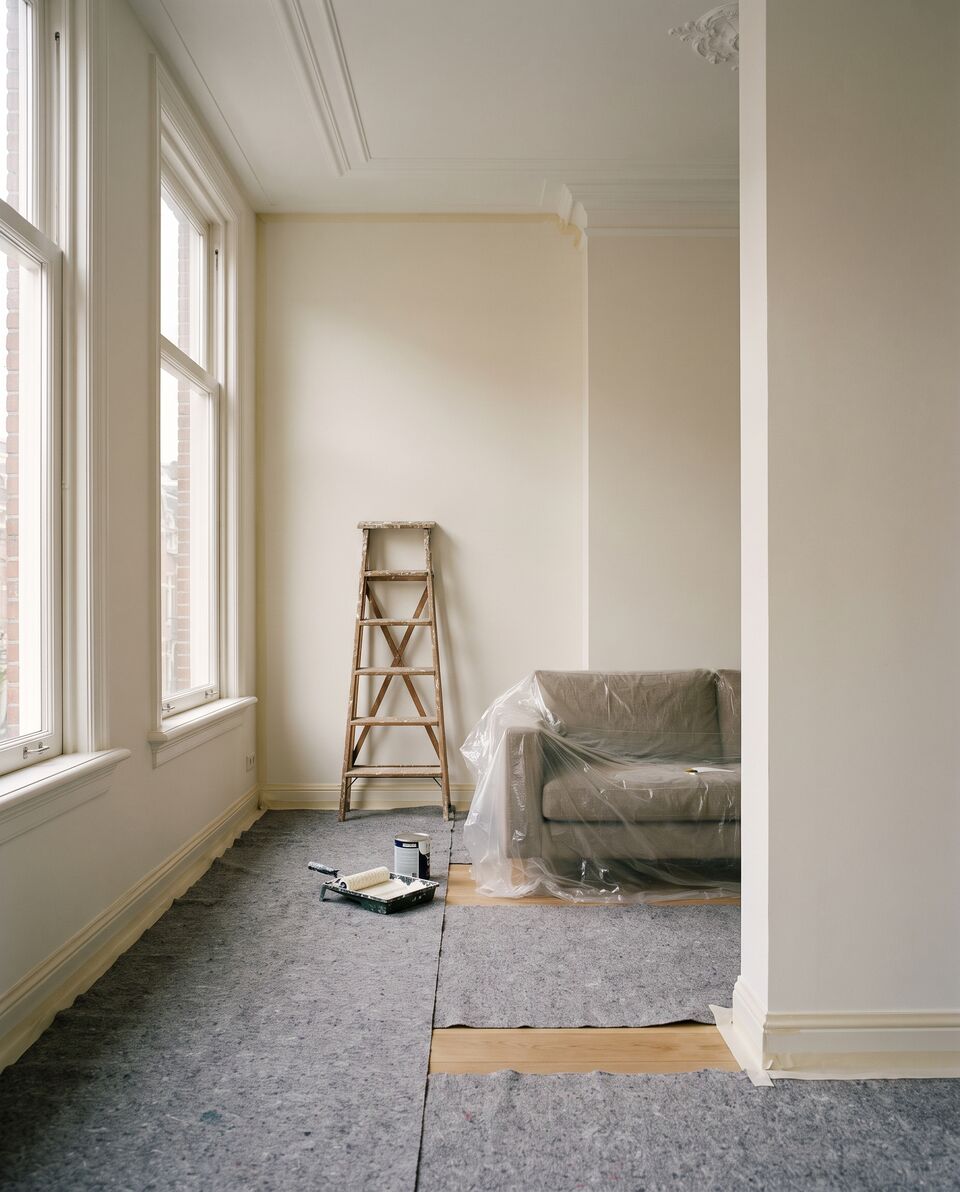 Woonkamer in een Amsterdamse bovenwoning tijdens het schilderwerk: vloer afgedekt met vlies, plinten afgeplakt, een trapje en een verfroller, de bank onder folie
