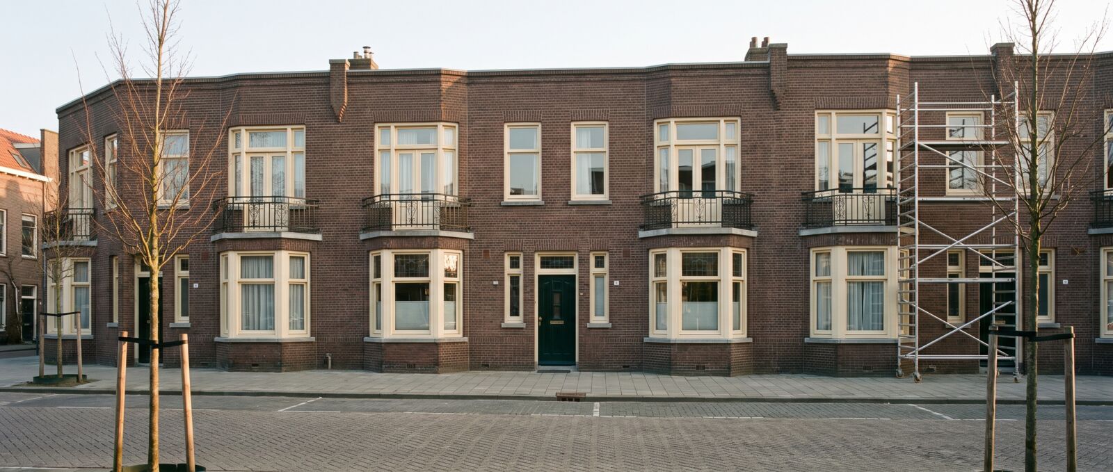 Rij Amsterdamse woningen uit de jaren twintig met crèmewitte kozijnen, een donkergroene voordeur en een rolsteiger aan het eind van de rij
