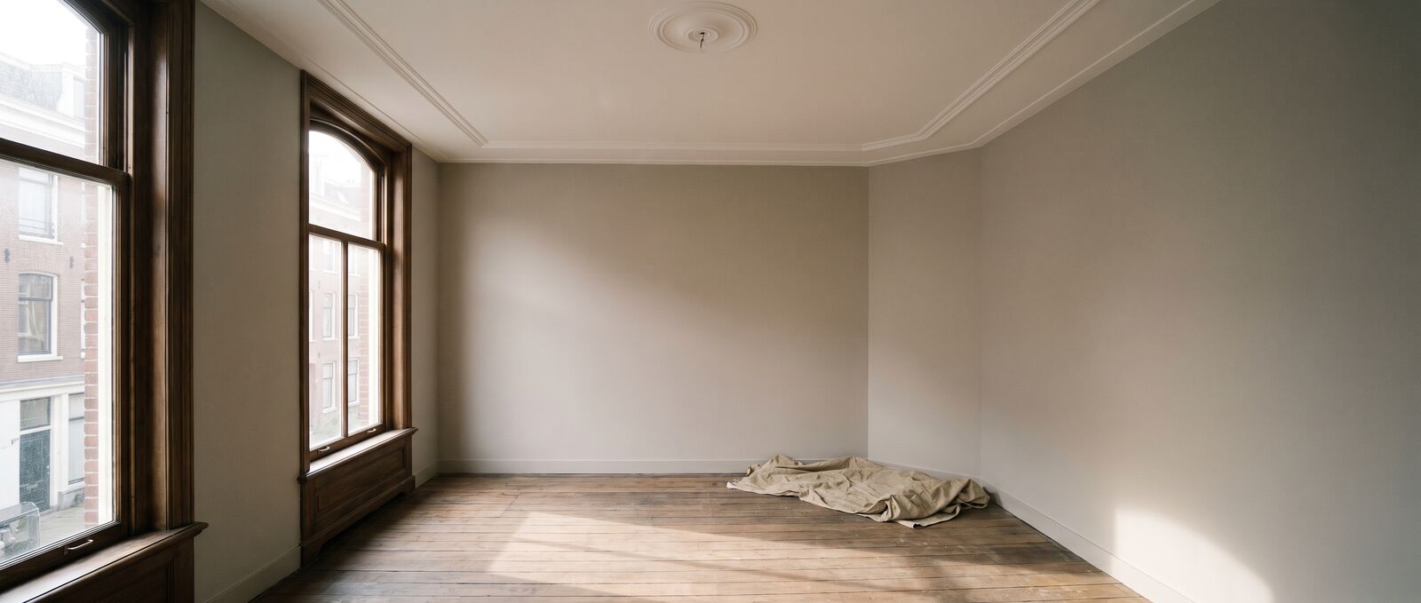 Lege kamer in een negentiende-eeuws Amsterdams huis met een hoog, egaal wit plafond met ornamentlijst en warmgrijze wanden in middaglicht